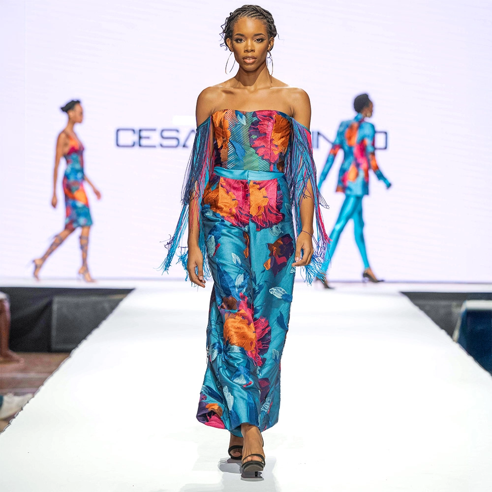 A model walks down a New York Fashion week runway in a Caribbean color influenced blouse and matching pants. The ensemble was manufactured by Zouhir NYC Group.